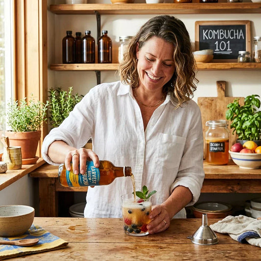 40+ year old woman pouring a raw kombucha into a glass while standing in her kitchen.