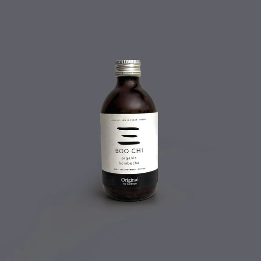 A bottle of BOO CHI Organic Kombucha on a solid dark gray background. The bottle is made of dark amber glass, has a silver metal screw cap, and features a minimalist white label with black text and graphic elements.