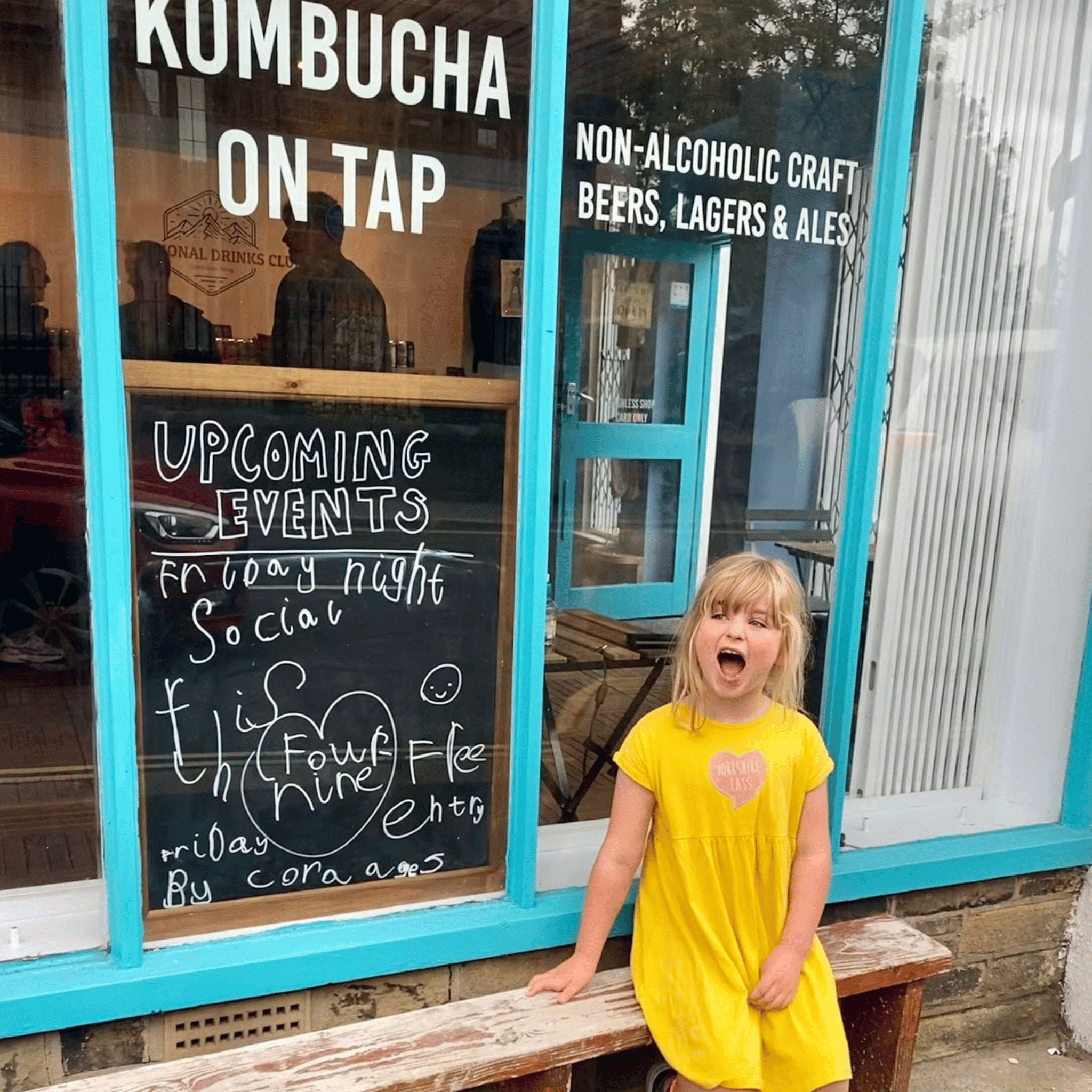 Child in a yellow dress standing in front of a store with a chalkboard sign and blue frame.