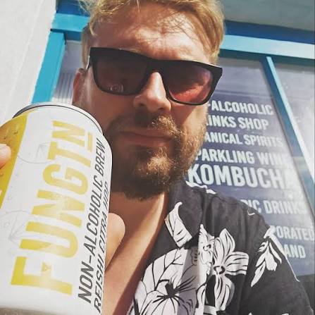 A man wearing black sunglasses and a black-and-white floral shirt holds up a can of Fungtm non-alcoholic brew in front of a storefront window with blue trim.