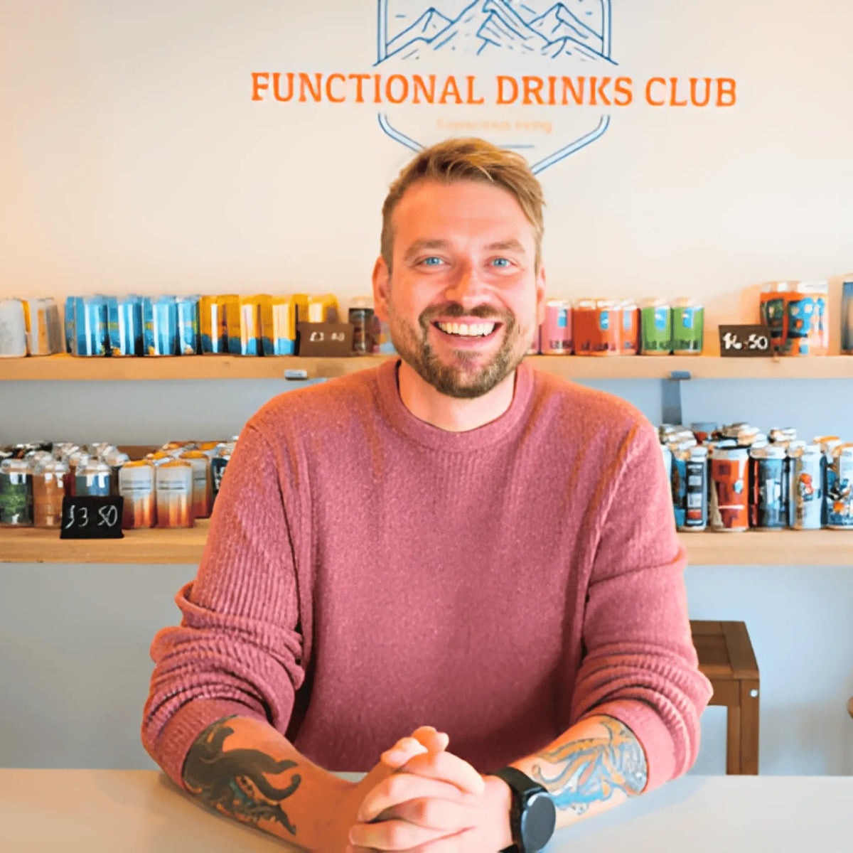 A smiling man with tattoos sits behind a counter in a shop, wearing a dusty rose knit sweater. Shelves behind him display numerous colorful canned drinks, and the wall features the logo and text 'FUNCTIONAL DRINKS CLUB'.