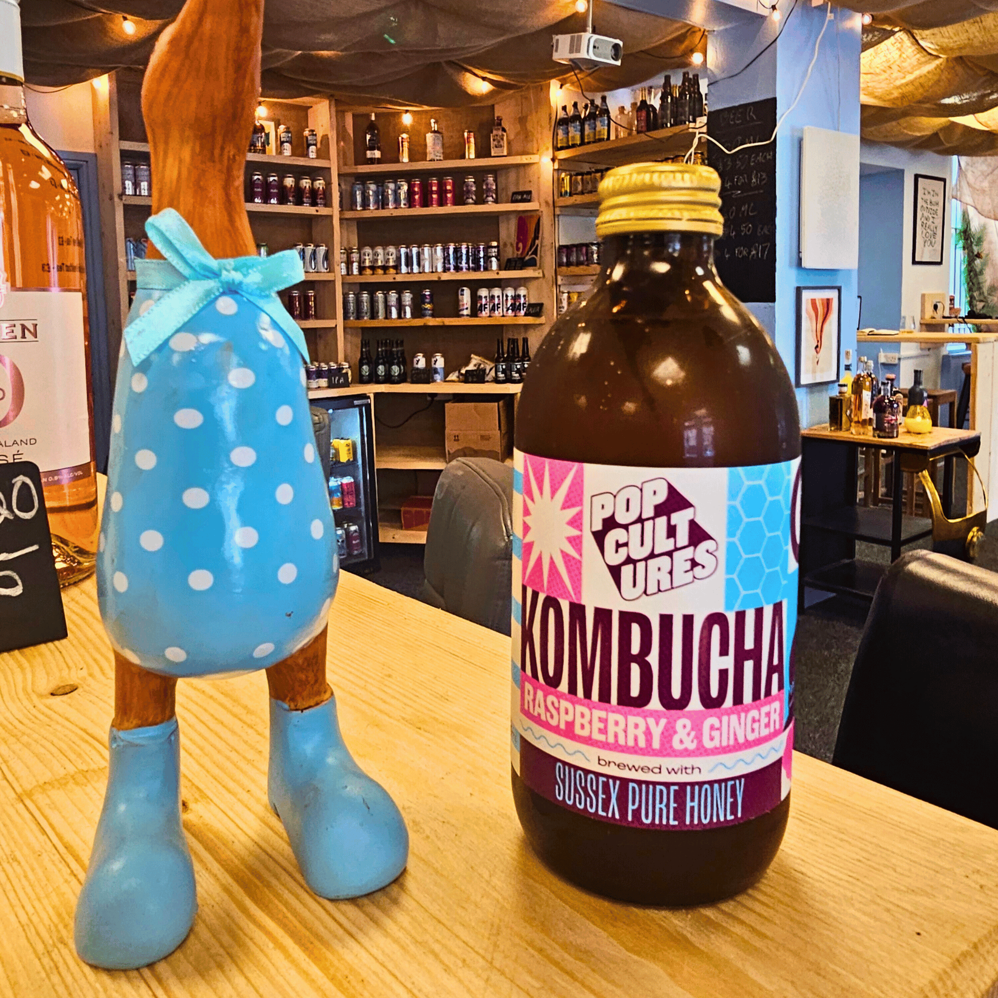 Pop Cultures kombucha bottle on a bar counter with a decorative bottle character in the background.