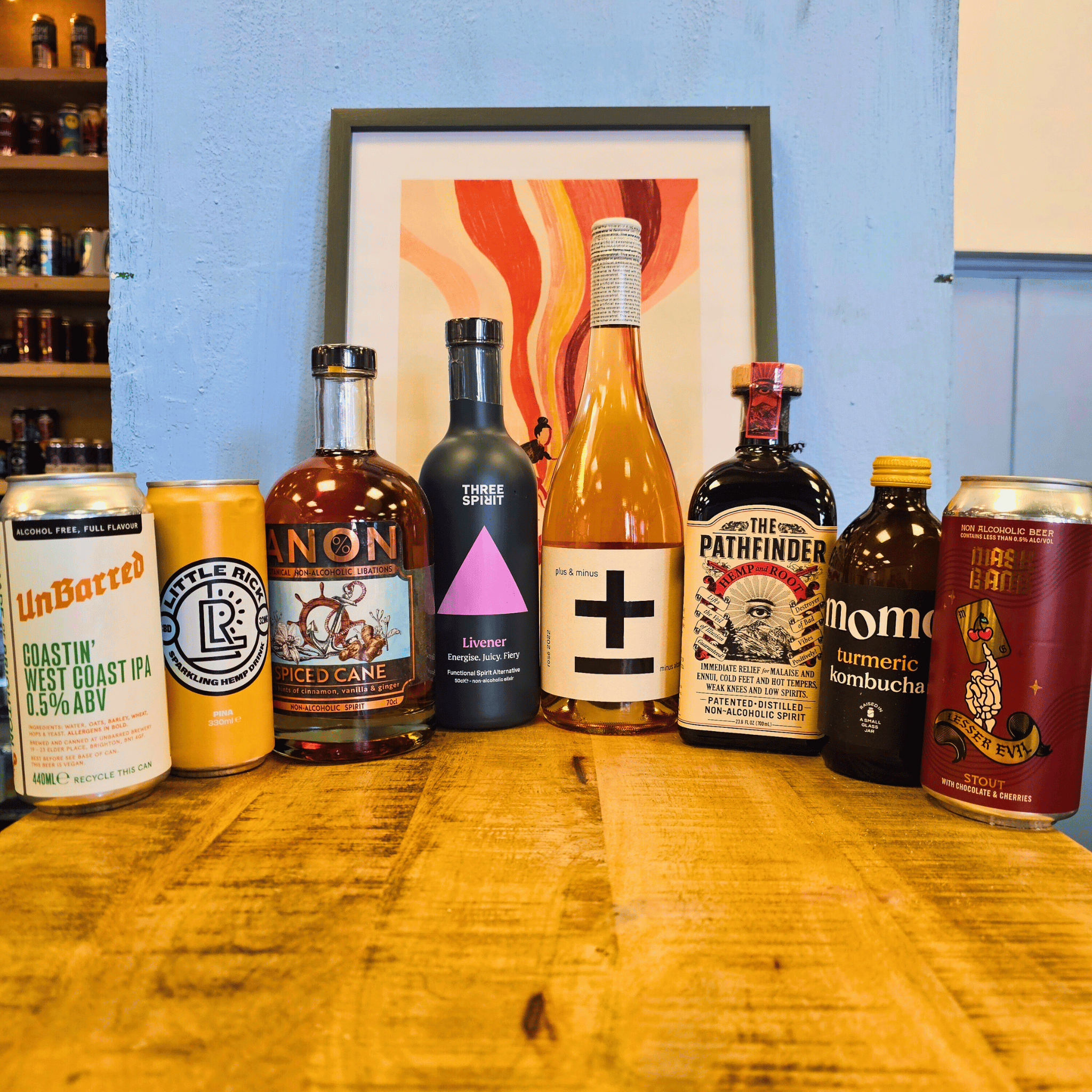 Collection of various bottles and cans on a wooden surface with a colorful framed picture in the background.