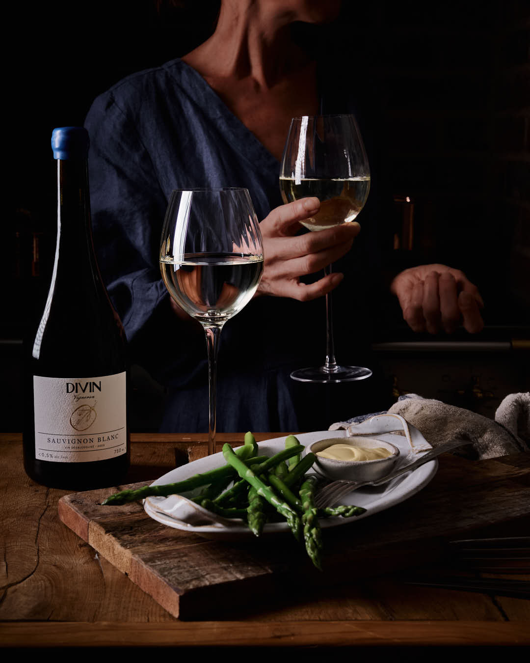 Person holding two wine glasses with a bottle of Divin wine and a plate of food on a wooden table.