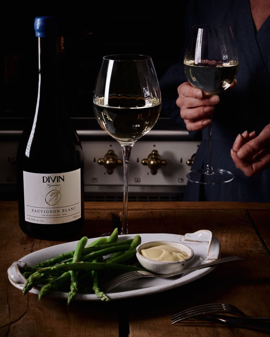 Bottle of Divin Sauvignon Blanc with two wine glasses and a plate of asparagus on a wooden table.