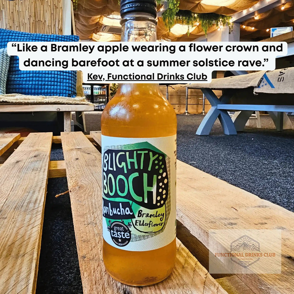 A bottle of Blighty Booch Kombucha with Bramley apple flavor sits on a wooden table in a rustic outdoor bar or cafe setting. A white text bubble at the top quotes Kev from Functional Drinks Club.
