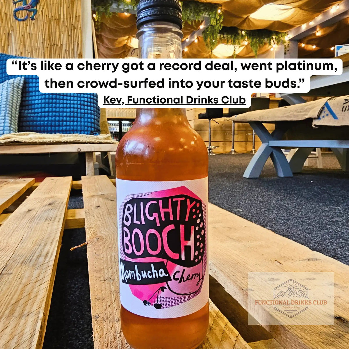 A bottle of Blighty Booch Kombucha Cherry drink placed on a wooden table in a cafe or restaurant setting, with a customer testimonial quote overlaid at the top.