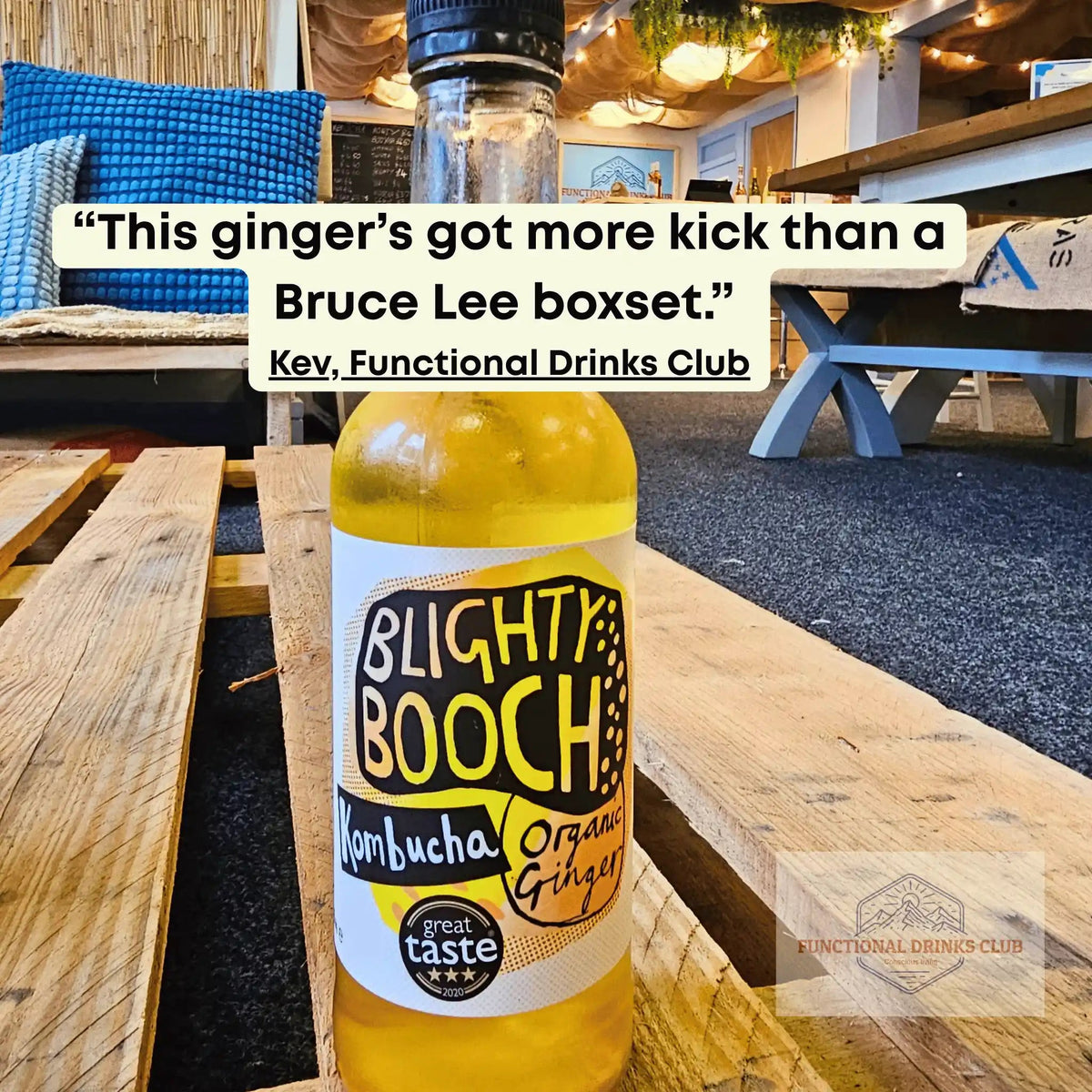 A bottle of Blighty Booch Kombucha Organic Ginger drink with a yellow liquid, set on a wooden table in a cafe environment. A quote overlay is visible at the top.