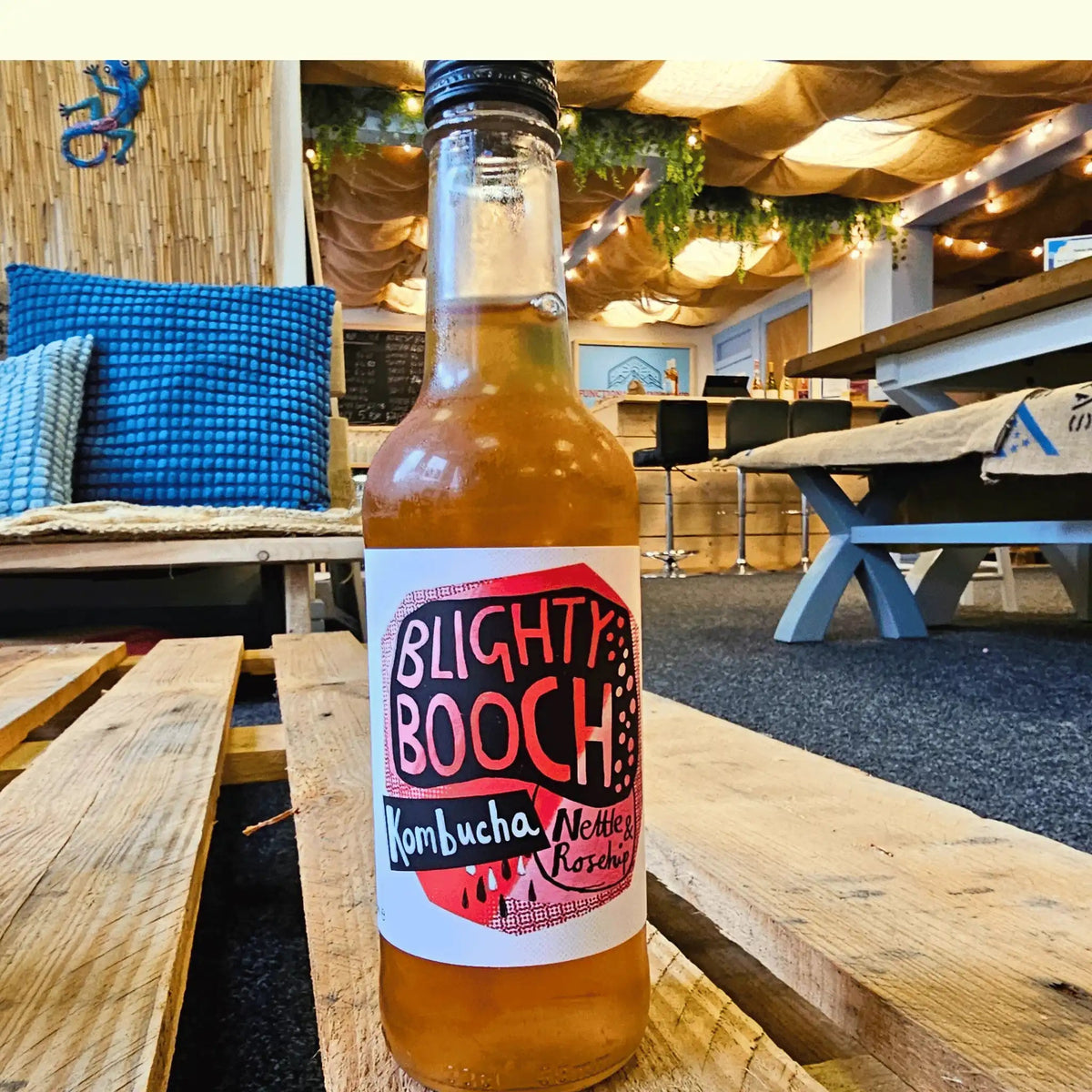 A bottle of Blighty Booch Kombucha with Nettle & Raspberry flavor, filled with amber-colored liquid, set on a wooden table in a rustic, cozy cafe environment.