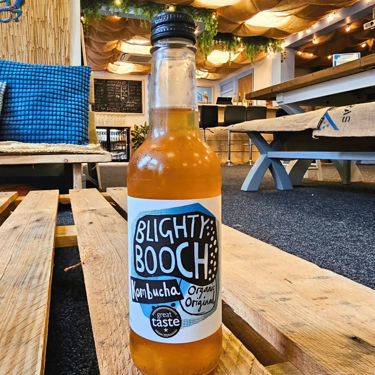 A bottle of Blighty Booch Organic Original Kombucha sits on a wooden table in a cafe setting.