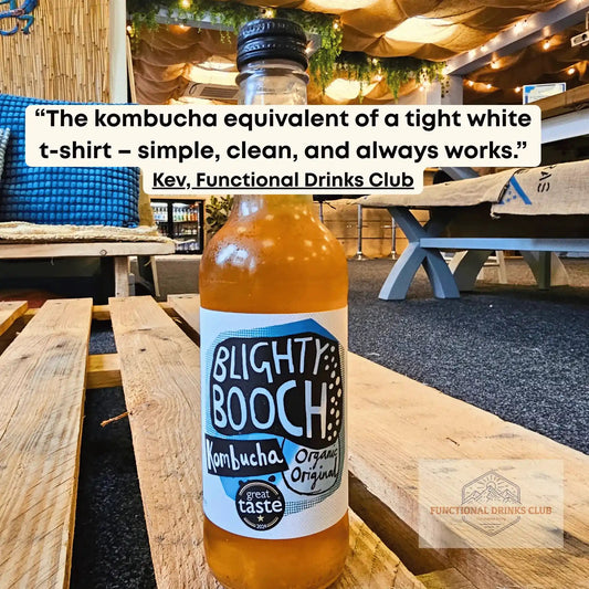 A bottle of  'Blighty Booch' kombucha organic original flavor, placed on a wooden pallet in an outdoor patio setting. A white text bubble with a quote is overlaid at the top, and a brand logo watermark is visible in the bottom right corner.