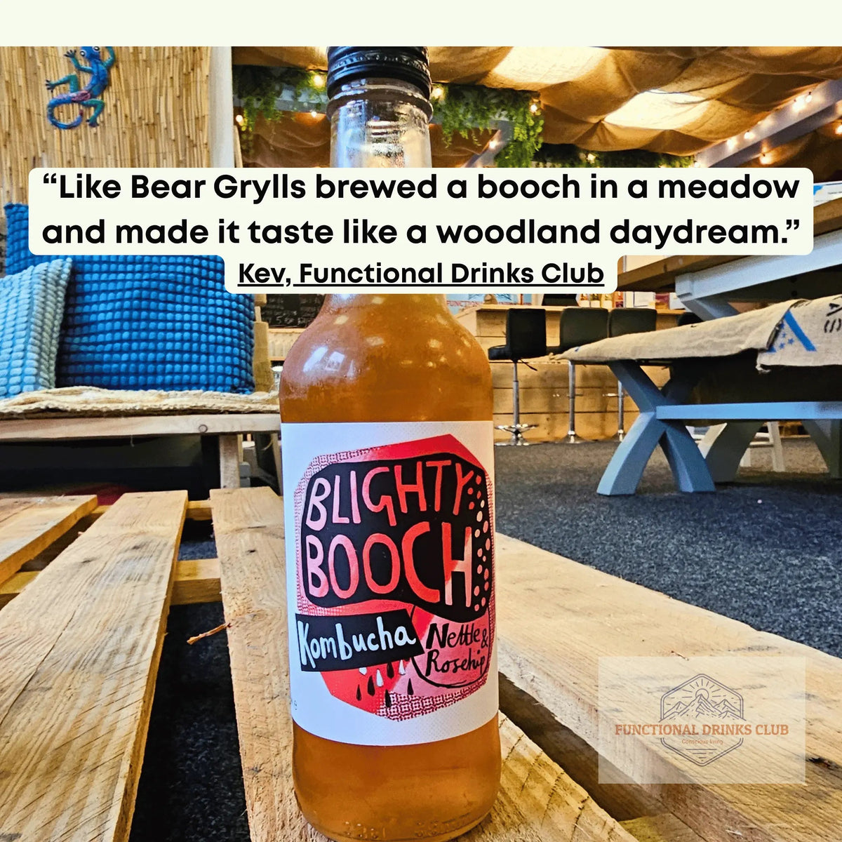 A bottle of Blighty Booch Kombucha Nectar Raspberry flavor sits on a wooden table in a rustic cafe setting. The bottle is filled with amber-colored kombucha and features a white label with red and black lettering.