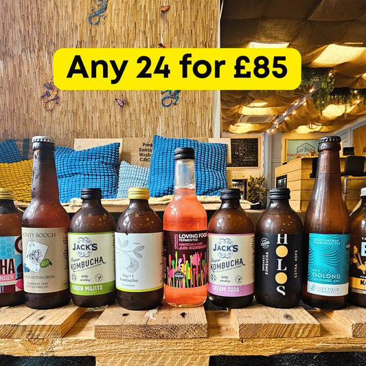 A promotional display of kombucha bottles arranged on wooden crates in a cozy indoor setting with a woven wall and blue pillows in the background. A large yellow banner at the top reads 'Any 24 for £85'.