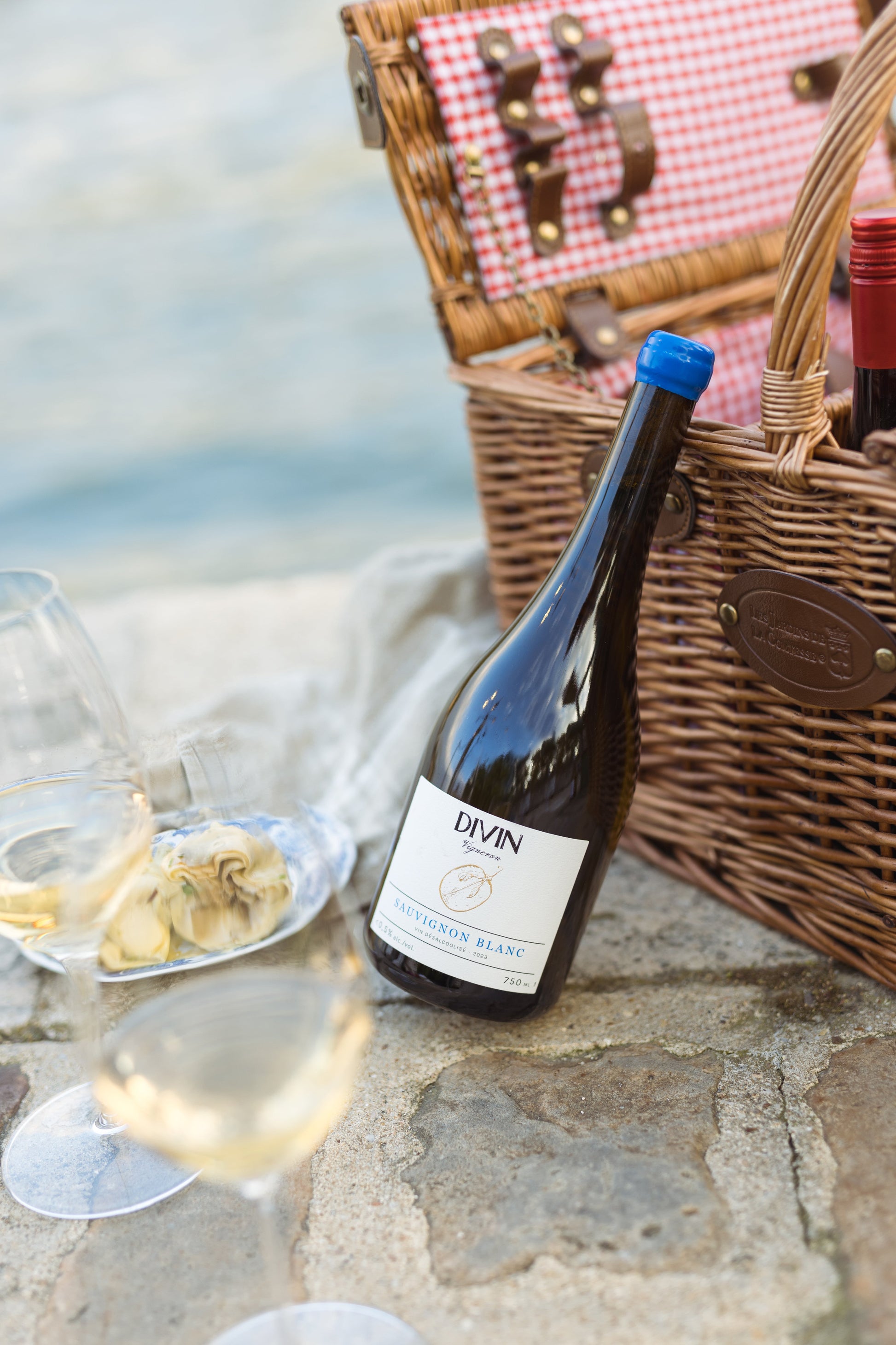 Bottle of Divin wine with a picnic basket and food on a stone surface by the sea.