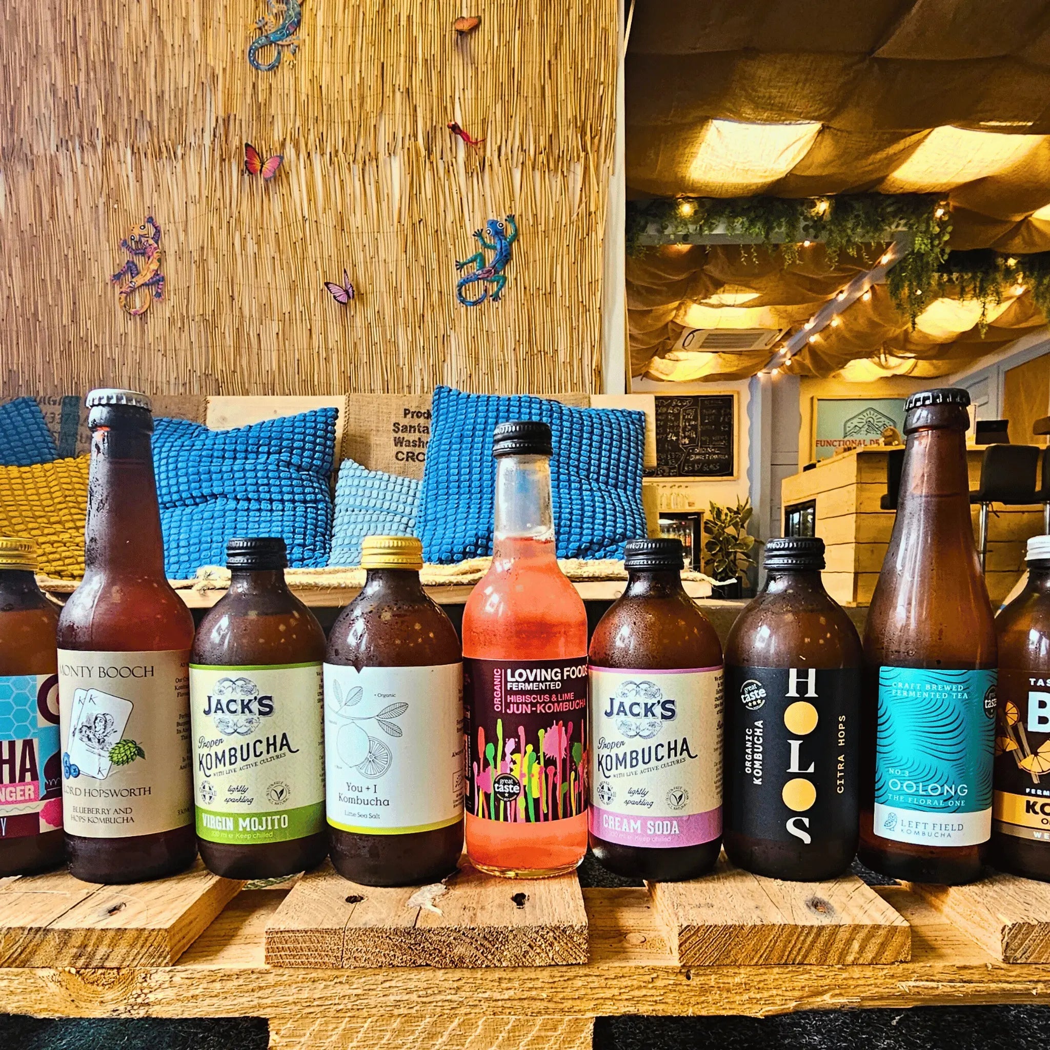 A row of kombucha bottles displayed on wooden crates in a cozy, rustic indoor setting with a woven wall backdrop and blue pillows at Functional Drinks Club in Otley.