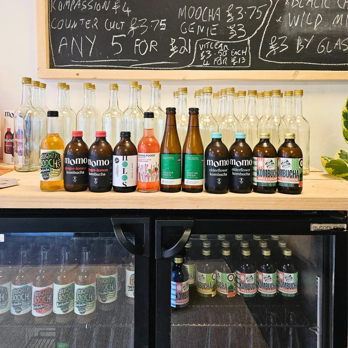 A lineup of different Kombucha bottles displayed on a wooden counter above a refrigerator, with a chalkboard menu in the background at Functional Drinks Club in Otley