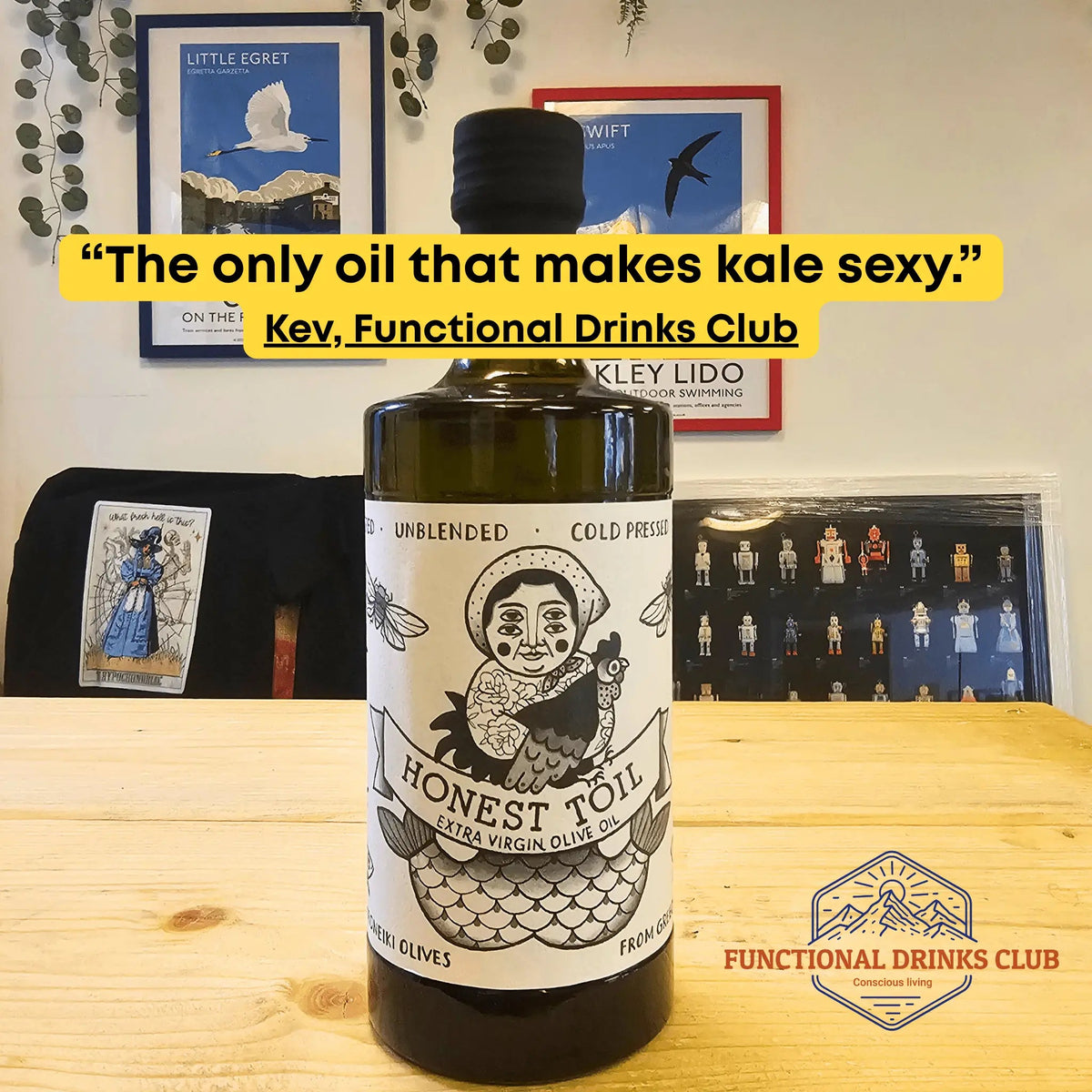 A bottle of Honest Toil Cold Pressed Unblended Olive Oil stands on a light wood table in a home setting, with framed posters on the wall behind it. A yellow text banner overlays the top portion of the image.