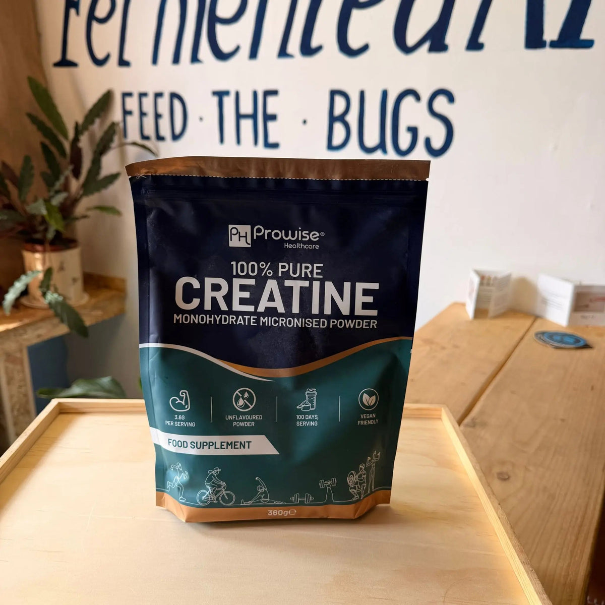 A stand-up pouch of Prowise Creatine 100% Pure Creatine Monohydrate Micronised Powder, placed on a light wooden tray in a home or studio setting with a white wall and blue lettering in the background.