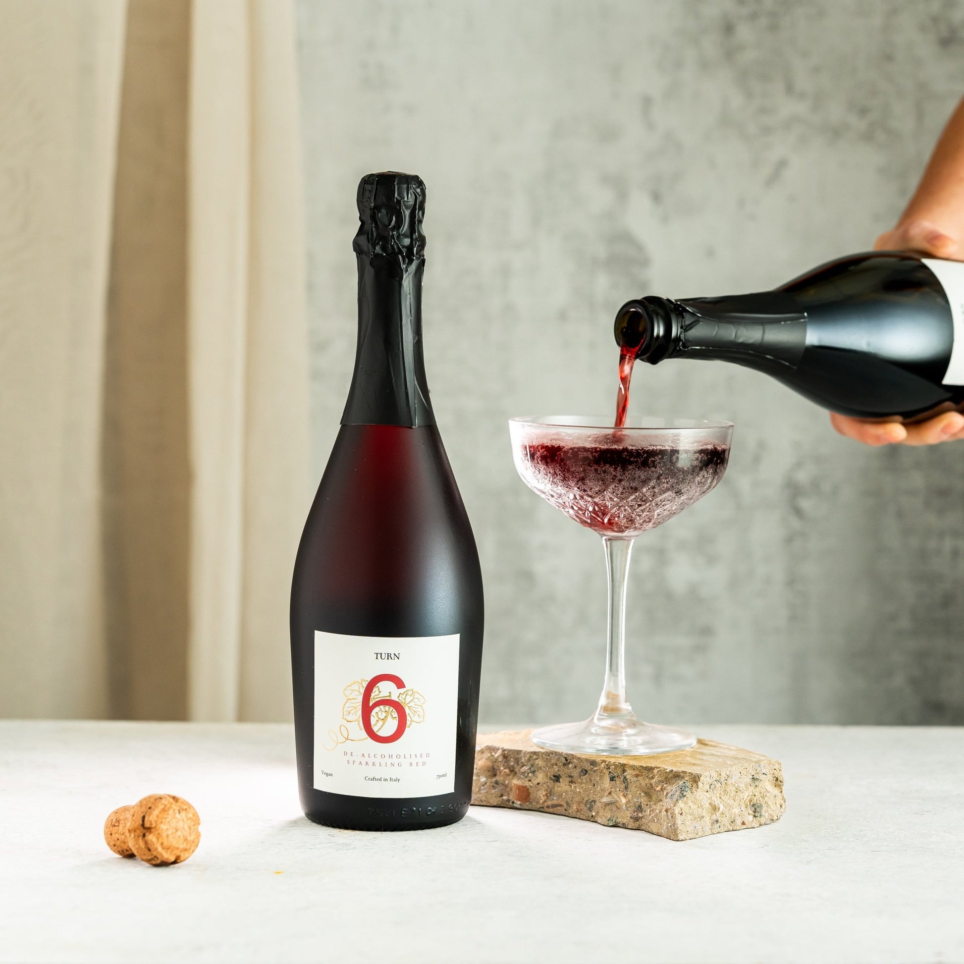 Bottle of red wine being poured into a glass with a cork on a neutral background