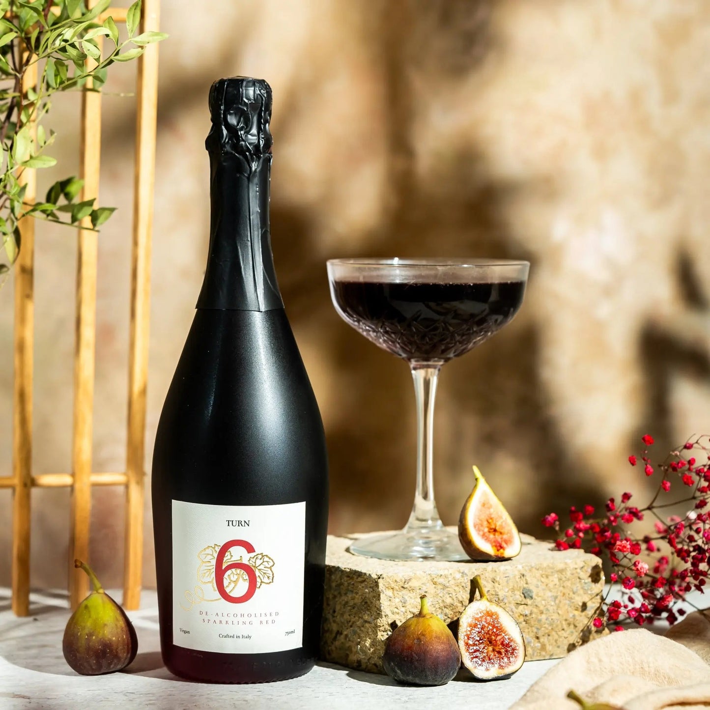 A bottle of TUN 6 years old sparkling wine stands next to a coupe glass filled with the dark red wine, styled with fresh figs and dried red flowers on a stone pedestal against a textured beige wall.