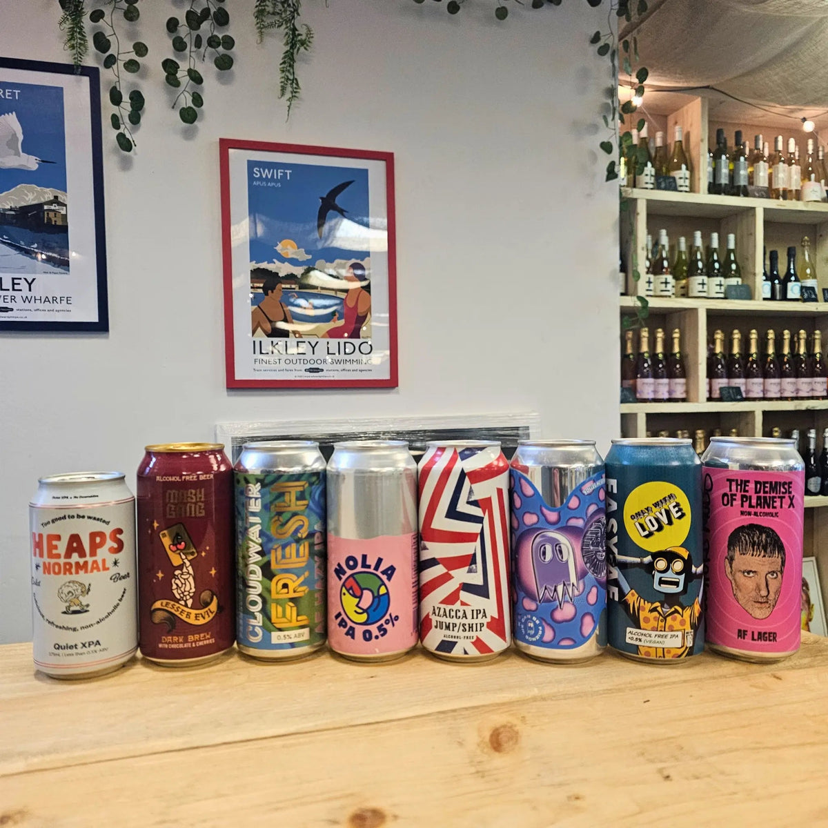 A row of eight craft beer cans in various colorful designs lined up on a light wooden counter in a shop setting with shelves of wine bottles and framed posters on the wall behind them.
