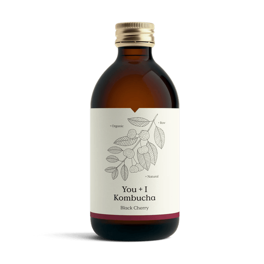 You + I Black Cherry raw kombucha in an amber glass bottle with a gold cap and a cream label with a botanical line drawing and a deep red band.